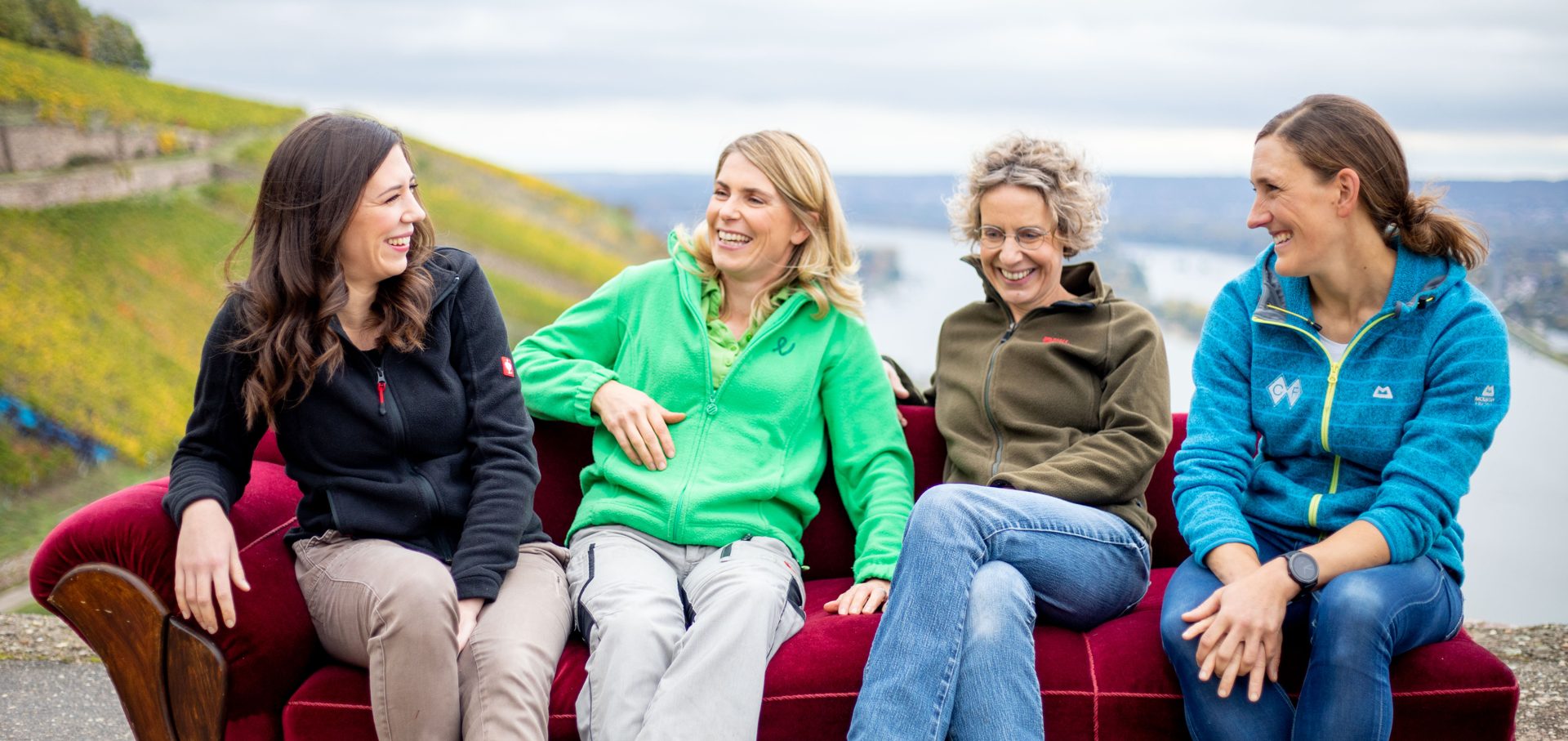 Vier lachende Frauen sitzen auf einem roten Sofa vor Weinbergen und dem Rhein